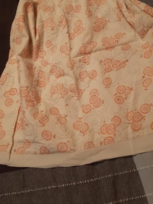 Blommig kjol med pärldetaljer - Söt kjol i ljusbeige med orange blommigt mönster och diskreta pärldetaljer. Kjolen har en lätt A-linjeform och är perfekt för dig som gillar romantiska och feminina plagg. Materialet känns mjukt och har en fin struktur. Storlek XXL