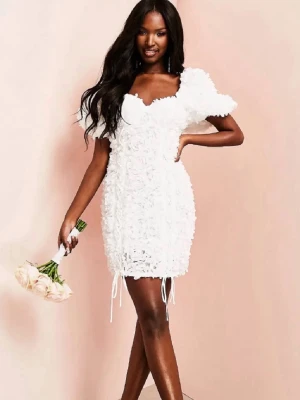 Vit kort klänning - Säljer en vit kort klänning med volangdetaljer och snörning längst ner. Klänningen har en v-ringad hals och puffiga korta ärmar. Lite spets/blomdetaljer över hela tyget. Perfekt till konfirmation och student!