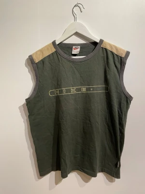 Ärmlös grön Nike t-shirt - Ärmlös t-shirt från Nike i mörkgrönt med beige axelpartier och grå kant runt hals och ärmhål. Framtill finns ett gult Nike-tryck. Tillverkad i mjuk bomull.
