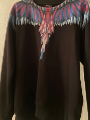 Marcelo burlon - Säljer en svart sweatshirt från Marcelo Burlon med ett färgglatt vingmönster i blått, rött och vitt över axlar och bröst. Tröjan har rund halsringning och långa ärmar. Perfekt för dig som vill sticka ut med en unik design. Passar även L 