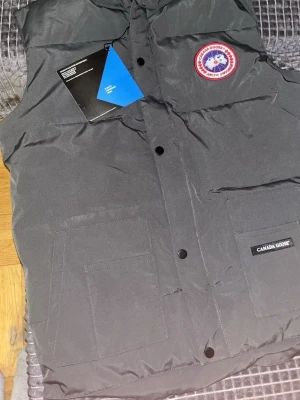 Svart dunväst från Canada Goose - Snygg svart dunväst från Canada Goose med klassisk logotyp på bröstet och två stora fickor framtill. Västen har tryckknappar och en stilren design, perfekt för lager-på-lager. Märkeslapp på ena fickan och hög krage för extra värme.