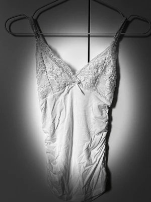 Vitt linne  - Söt vitt linne med smala axelband och v-ringad spets upptill. Dekorativ liten rosett framtill och mjukt, stretchigt tyg nedtill. ✨