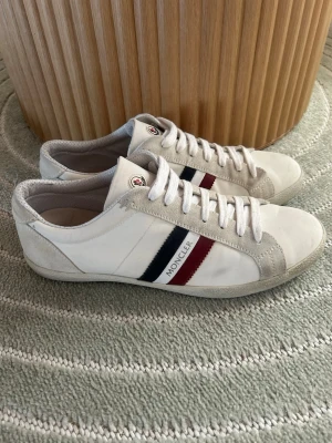 Vita Moncler sneakers med ränder - Snygga vita sneakers från Moncler med klassiska röda och marinblå ränder på sidan. Skorna har vita skosnören, beige mockadetaljer och Moncler-logga på plösen. Tillverkade i läder och mocka för en clean och stilren look.