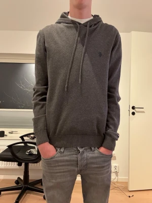 Mörkgrå hoodie från U.S. Polo Assn. - Mörkgrå hoodie från U.S. Polo Assn. med dragsko i huvan och diskret logga på bröstet. Tillverkad i mjukt material med ribbade muddar vid ärmslut och nederkant. Perfekt för en chill och stilren look.