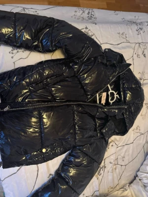 Glansig svart pufferjacka - Snygg svart pufferjacka med glansig finish och dragkedja framtill. Jackan har hög krage och quiltad design som ger en cool look. Perfekt för kalla dagar när du vill hålla värmen och ändå se trendig ut. Ordinarie pris va 800. 
