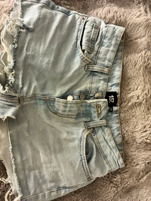 Ljusblå jeansshorts från 157 - Säljer ett par ljusblå jeansshorts från 157 med råa, fransiga kanter och klassiska femficksdetaljer. Shortsen har knappar framtill och är midwaist. Perfekta för sommarens alla äventyr!