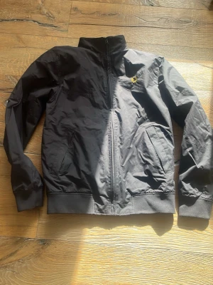 Svart bomberjacka från Fred Perry - Svart bomberjacka från Fred Perry med guldfärgad logga på bröstet och gul rand på insidan av kragen. Jackan har dragkedja framtill, två snedställda fickor och ribbade muddar vid ärmslut och midja. Perfekt för en stilren och sportig look.