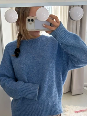 Blå stickad tröja från Zara  - Mysig blå stickad tröja med oversized passform och breda ribbade muddar vid ärmslut och nederkant. Tröjan har rund halsringning och är perfekt för lager-på-lager-stil. Materialet ser ut att vara mjukt och fluffigt, vilket ger en avslappnad vibe.