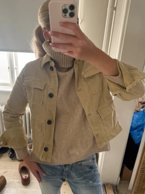 Beige overshirt med fickor - Stilren beige overshirt med fyra stora fickor framtill och svarta knappar. Modellen är kortare och har klassisk krage. Perfekt att slänga över en stickad tröja för en avslappnad look. Materialet ser ut att vara bomull eller en bomullsblandning.
