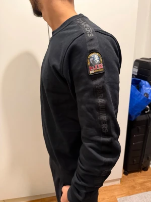 Svart sweatshirt från Parajumpers helt ny - Cool svart sweatshirt från Parajumpers med logotyp på ärmen och text längs axeln. Tröjan har rund halsringning, ribbade muddar och är tillverkad i mjukt material. Perfekt för dig som gillar stilrena och sportiga plagg.