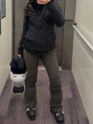 Mörkgröna täckbyxor från J.Lindeberg - Snygga mörkgröna täckbyxor från J.Lindeberg med dragkedjeficka och bälteshällor. Byxorna har en slimmad passform och är tillverkade i ett stretchigt, vattenavvisande material som passar perfekt för skidåkning eller vinteraktiviteter. Diskret JL-logga på benet.