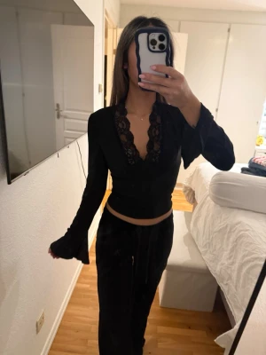Svart croppad topp med spets - Säljer en svart croppad topp med långärmad design och djup v-ringning med snygga spetsdetaljer. Perfekt för dig som gillar en stilren look med lite edge. Passformen är tight och materialet känns mjukt mot huden. 