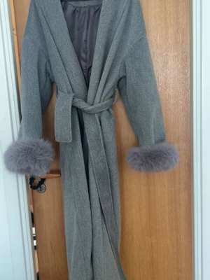 Grå kappa med päls  - Lång grå kappa med stora slag, knytskärp i midjan och fluffiga pälsdetaljer vid ärmsluten. Kappan har en mjuk och elegant look, perfekt för att höja din outfit under kyliga dagar. 