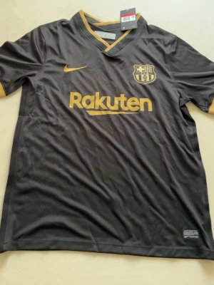 Svart FC Barcelona matchtröja Nike - Snygg svart FC Barcelona matchtröja från Nike med guldiga detaljer och loggor. Tröjan har V-ringning, korta ärmar och tryckta sponsorer som Rakuten och Unicef. Tillverkad i lätt och ventilerande funktionsmaterial, perfekt för fotboll eller träning.