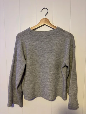 Grå stickad tröja i ullmix - Mysig grå stickad tröja med rund hals och långa ärmar. Tröjan har en avslappnad passform och ribbstickade muddar vid ärmslut och nederkant. Perfekt för kyliga dagar och enkel att matcha med olika outfits.