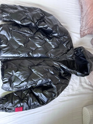 Svart glansig pufferjacka från Fendi - Pris kan diskuteras. Har haft den ett tag bara och den är helt ny. Inga skador eller något sånt. Säljer en svart glansig pufferjacka från Armani med huva och dragkedja framtill. Jackan har Armani-loggor i quiltat mönster över hela ytan och en röd detalj på ärmen. Perfekt för kalla vinterdagar och riktigt snygg streetstil.