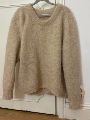 Beige fluffig stickad tröja Wera - oversize och mysig beige stickad tröja från Wera med rund halsringning och långa ärmar. Tröjan är fluffig och har ribbstickade muddar vid ärmslut och hals. Perfekt för lager på lager och en avslappnad look.