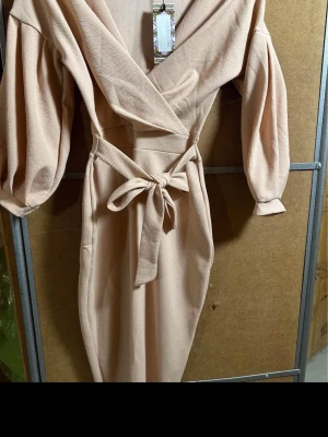 Beige omlottklänning från Back Up - Snygg beige omlottklänning från Back Up med puffärmar och knytskärp i midjan. Djup v-ringning och struktur i tyget ger en trendig look. Perfekt för dig som gillar stilrena och moderna plagg.