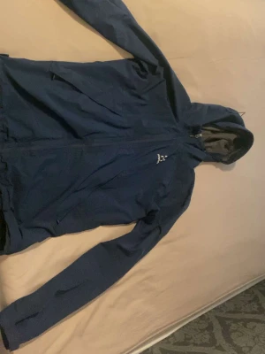 Blå vindjacka från Craft med huva - Snygg blå vindjacka från Haglöfs med hel dragkedja, justerbar huva och två sidofickor med dragkedja. Jackan har en sportig look och är tillverkad i lätt, vindtätt material som passar perfekt för utomhusaktiviteter.