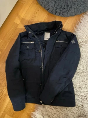 Mörkblå jacka från J.Lindeberg - Riktigt fin mörkblå jacka från J.Lindeberg med dragkedja och knappar framtill. Jackan har flera fickor med både knapp- och dragkedjedetaljer samt en hög krage. Perfekt och stilren nu inför våren.