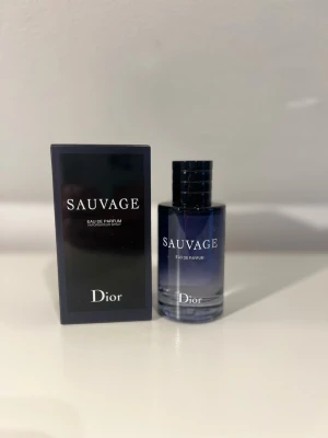 Dior Sauvage Eau de Parfum - Dior Sauvage Eau de Parfum kommer i en elegant mörkblå glasflaska med svart lock och vit text. Doften är maskulin och modern, och flaskan har en stilren, rund form. Förpackningen är svart med tydlig logga och produktnamn i vitt.