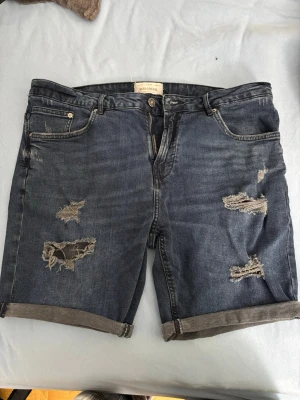 Mörkblå slitna jeansshorts Pull&Bear - Säljer ett par mörkblå jeansshorts från Pull&Bear med slitna detaljer och upprullade benslut. Shortsen har fem fickor och klassisk knappgylf. Perfekta för en avslappnad och trendig stil.