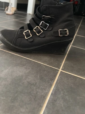 Svarta kilklackade stövletter med spännen - Snygga svarta stövletter med kilklack och fyra dekorativa spännen i silver. Skorna har en cool streetstil och är tillverkade i tygmaterial. Perfekta för dig som vill ha en edgy look med lite extra höjd.