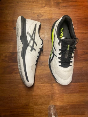 Vita Asics sneakers med svarta detaljer - Snygga vita sneakers från Asics med svarta och neongula detaljer. Skorna har en låg profil, svart snörning och Asics-logga på sidan. Ovandelen är i mesh och syntetmaterial för bra andningsförmåga och komfort. Perfekta för en sportig och stilren look.