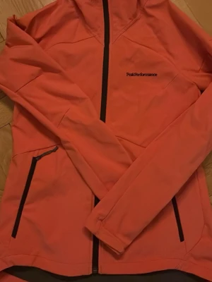 Rosa vindjacka från Peak Performance - Sportig rosa vindjacka från Peak Performance med svarta dragkedjor och hög krage. Jackan har lång ärm och två fickor framtill. Perfekt för dig som gillar en aktiv stil och vill ha en snygg jacka till vardags.