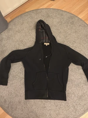Svart hoodie från Burberry - Snygg svart hoodie från Burberry med dragkedja och klassiskt rutigt foder i huvan. Broderad Burberry-logga på bröstet och två stora fickor framtill. Perfekt för en stilren och avslappnad look.Den har ingen dragkedja men det är lätt att fixa men annars funkar den perfekt!!!