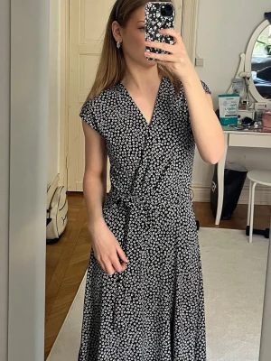 Svart blommig omlottklänning - Svart långklänning med vita små blommor och omlott-design. Klänningen har korta ärmar, v-ringning och knytband i midjan som ger en snygg passform. Perfekt för dig som gillar en feminin och stilren look.