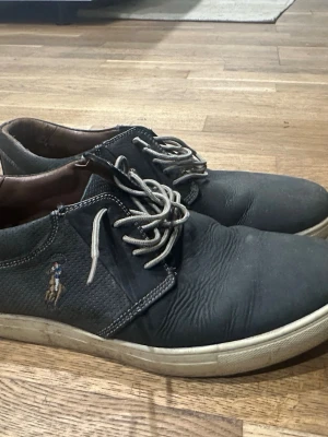 Svarta loafers från Polo Ralph Lauren - Säljer ett par svarta loafers från Polo Ralph Lauren med klassisk broderad logga på sidan. Skorna har vita sulor och grå snören, samt detaljer i läder och textil. Perfekta för en avslappnad men ändå stilren look.