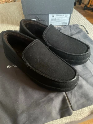 Svarta tofflor från Ermenegildo Zegna - Stilrena svarta tofflor från Ermenegildo Zegna i merinoull med mjukt foder. Klassisk slip-on modell med rundad tå och diskreta sömmar. Perfekta för att chilla hemma och hålla fötterna varma. Kommer med originalpåse och box.