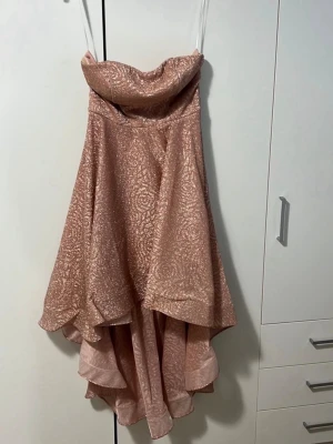 Glittrig axelbandslös rosa klänning - Säljer en glittrig axelbandslös klänning i rosa med volangig nederdel och asymmetrisk längd. Klänningen har en snygg omlottkänsla upptill och är perfekt för dig som vill sticka ut. Materialet känns lyxigt och skimrande.