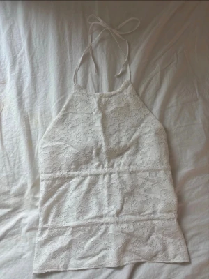 Vit spetsig halterneck linnetopp homemade - Säljer en vit halterneck linnetopp med spetsdetaljer och knyt i nacken och ryggen. Toppen har en ärmlös design och är perfekt för varma dagar. Materialet är lätt och luftigt med ett romantiskt spetsmönster. Homemade!