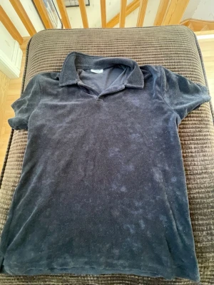 Mörkblå frottéskjorta från Kappahl - Mjuk och skön mörkblå skjorta från Kappahl i frottématerial. Skjortan har klassisk krage och korta ärmar, perfekt för en avslappnad och cool stil. Passar dig som gillar bekväma plagg med lite retrovibe.