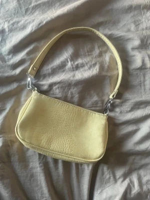 Ljusgul axelväska i krokopräglat skinn - Trendig ljusgul handväska med axelrem och krokodilpräglat mönster. Väskan har en stilren, rektangulär form och silverfärgade metalldetaljer. Perfekt accessoar för att lyfta din outfit.