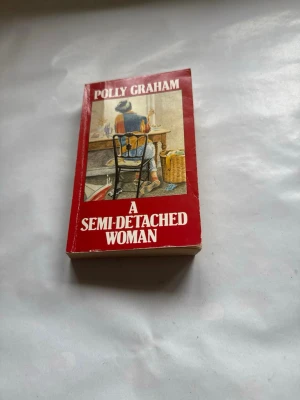 A Semi-Detached Woman Polly Graham - Engelskspråkig roman med röd och vit framsida. Omslaget visar en kvinna som sitter vid ett bord, illustrerat i mjuka färger. Pocketbok med klassisk design och tydlig titeltext. Perfekt för dig som gillar samtida skönlitteratur.