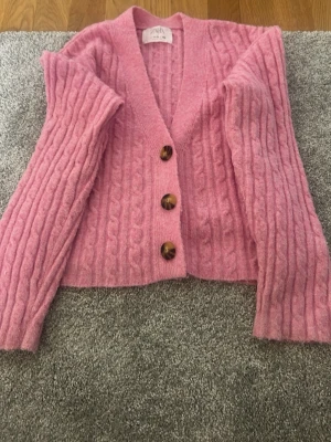 Rosa kabelstickad kofta från Zara - Säljer en rosa kabelstickad kofta från Zara med tre stora bruna knappar framtill. Koftan har v-ringning, lång ärm och är gjord i ett mjukt, fluffigt material. Perfekt för dig som gillar en mysig och trendig look.