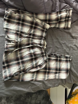 Rutiga pyjamasbyxor XS från lager 157 - Mysiga pyjamasbyxor från lager 157 i storlek XS. Byxorna har ett klassiskt rutigt mönster i svart, vitt och beige med dragsko i midjan och mjukt bomullstyg. Perfekta för chillkvällar hemma eller när du vill vara extra bekväm.