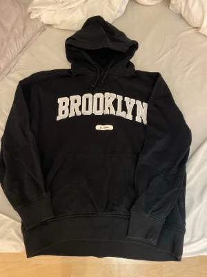 Svart hoodie med BROOKLYN-tryck - Svart hoodie med stor huva och dragsko, BROOKLYN-tryck i vitt på bröstet och texten EST.1984 under. Klassisk känguruficka framtill och ribbade muddar vid ärmar och nederkant. Perfekt för en avslappnad streetwear-look.