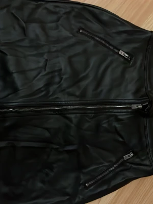 Svart skinnjacka med dragkedjor - Snygg svart skinnjacka med raka linjer och två snedställda dragkedjefickor framtill. Jackan har en klassisk look med en rejäl dragkedja i mitten och stilrena detaljer. Perfekt för dig som gillar en tidlös och cool stil. Köpt utomlands använd 3 gånger 