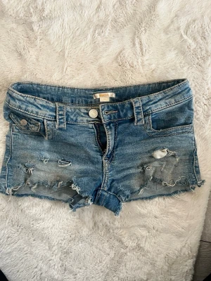 Blå slitna jeansshorts från Gina - Säljer ett par blå jeansshorts med slitna detaljer. Som man ser på bilden är dom lite smutsiga men det är bara jord, så det går bort i tvätten.