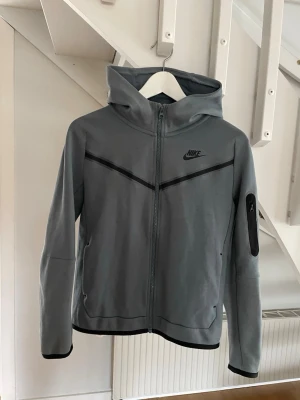 Grå Nike hoodie med dragkedja - Snygg grå hoodie från Nike med hel dragkedja framtill och svart Nike-logga på bröstet. Hoodien har svarta detaljer, huva och en praktisk ficka med dragkedja på ärmen. Tillverkad i mjukt och stretchigt material som känns skönt mot huden.