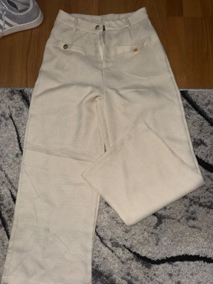 Beiga kostymbyxor med guldknappar - Snygga beiga kostymbyxor med raka ben och hög midja. Byxorna har dekorativa guldknappar vid fickorna och ett diskret vävt mönster i tyget. Perfekta för en stilren och trendig look. Materialet känns mjukt och har en lätt struktur. Storleken är petite XS så längden passar de under 160