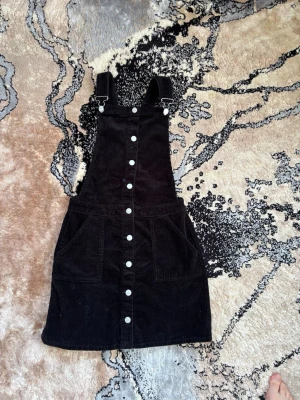 Svart manchester jumpsuit med knappar - Snygg svart jumpsuit i manchestertyg med justerbara axelband och knäppning framtill. Stora fickor framtill ger en cool och avslappnad look. Axelbanden korsas i ryggen för extra stilpoäng. Perfekt att matcha med en t-shirt eller tröja under. Anledningen till att jag säljer denna är på grund av att den inte passar min ålder längre. 