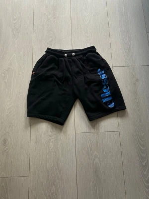 Svarta shorts från Ellesse med tryck - Svarta shorts från Ellesse med blått logotryck längs ena benet. Modellen har resårmidja med snörning och två sidofickor. Tillverkade i mjuk bomull, perfekta för sommaren.