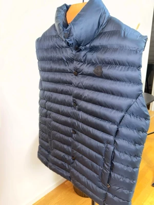 Mörkblå dunväst från Moncler - Snygg mörkblå dunväst från Moncler med hög krage och knappstängning framtill. Västen har ett diskret Moncler-märke på bröstet och är tillverkad i glansigt, quiltat material med två sidofickor. Perfekt för lager-på-lager och streetstyle.