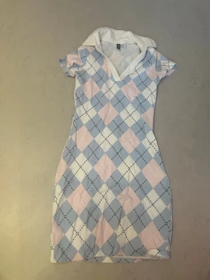 Pastellrutig klänning från Indeci - Kortärmad klänning från Indeci med pastellrutor i ljusblått, vitt och ljusrosa. Klänningen har en vit krage och v-ringning, samt ett rutigt mönster över hela plagget. Tillverkad i mjuk bomull, perfekt för en avslappnad och trendig look. Andvänt en gång 