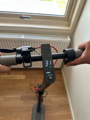 Svart elscooter med display - Säljer en svart elscooter med digital display som visar hastighet och batterinivå. Scootern har grå handtag, framlykta och röda detaljer på kablarna. Utrustad med handbroms och bred fotplatta för stabil körning.